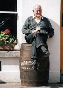Photo of older man sitting on top of whiskey barrel drinking stout