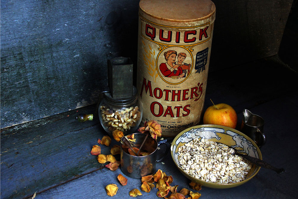 Photo of dry oats in ceramic bowl, antique nut grinder, dried roses, antique oats container, apple